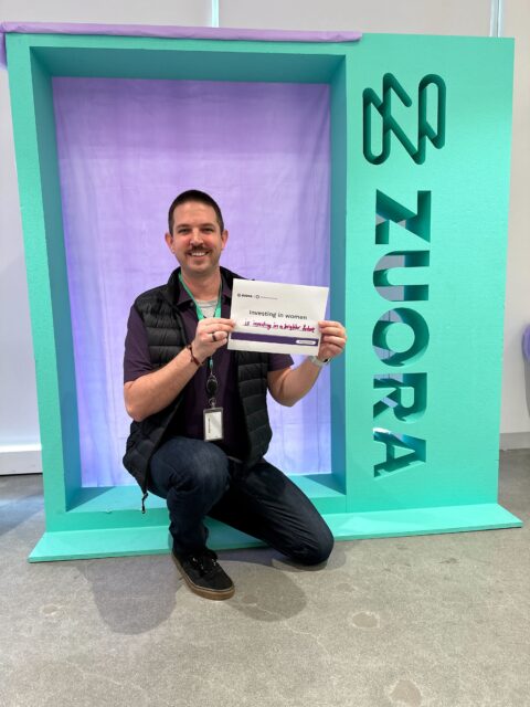 A man crouching in front of a purple wall with a check.