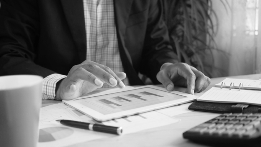 A black and white image of a person's hands reviewing charts on a digital tablet with papers, a notebook, and a calculator on the desk.