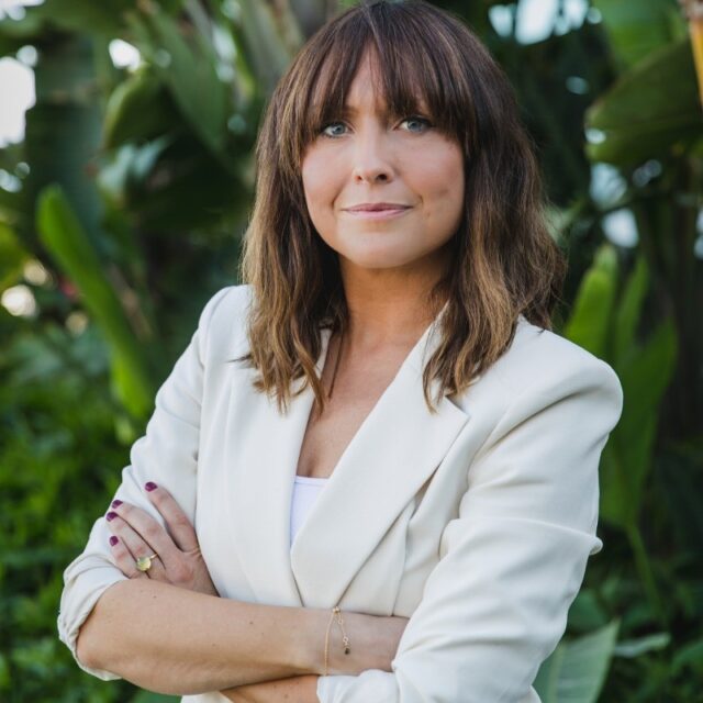 A woman in a white blazer standing in front of plants.