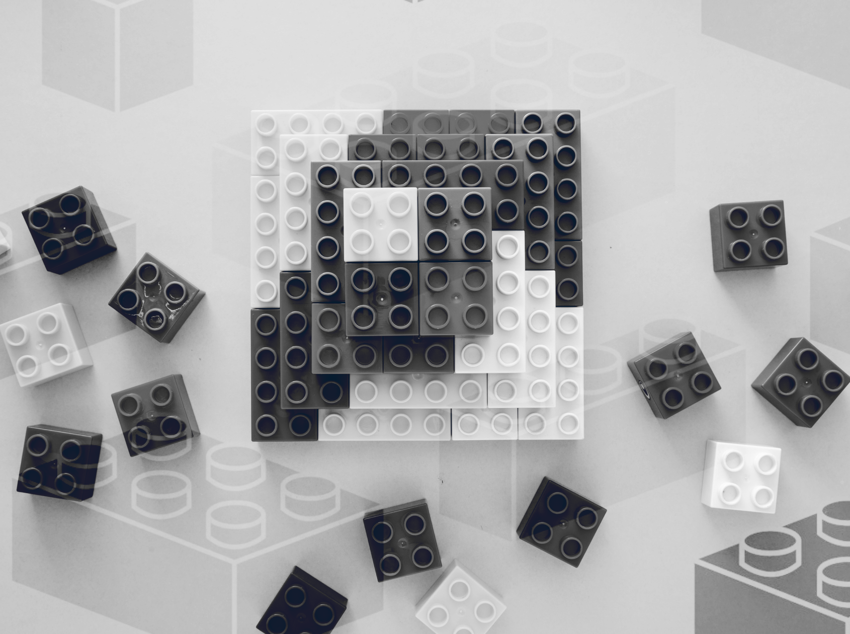 Monochrome image of various lego pieces scattered around a central square lego construction resembling the letter 'a'.