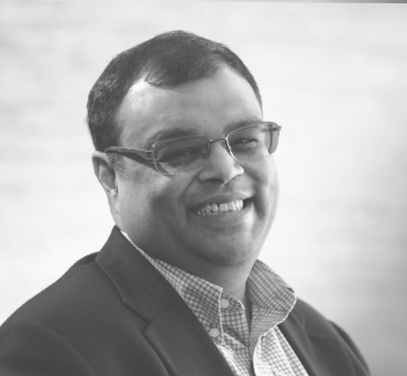 Black and white portrait of a smiling man wearing glasses and a plaid jacket.