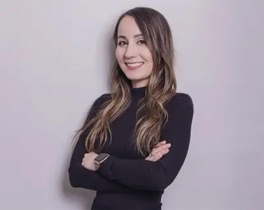 A woman with long, wavy brown hair wearing a black long-sleeve top and a smartwatch is smiling with her arms crossed against a plain light background, ready to explore exciting opportunities at Zuora careers.