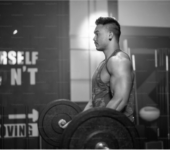 A muscular man lifting a barbell in a gym, focused and wearing a tank top, with motivational posters in the background.