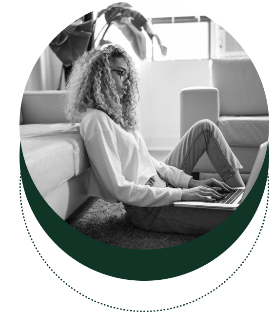 A person with curly hair wearing glasses is sitting on the floor, leaning against a couch, and working on an open laptop.