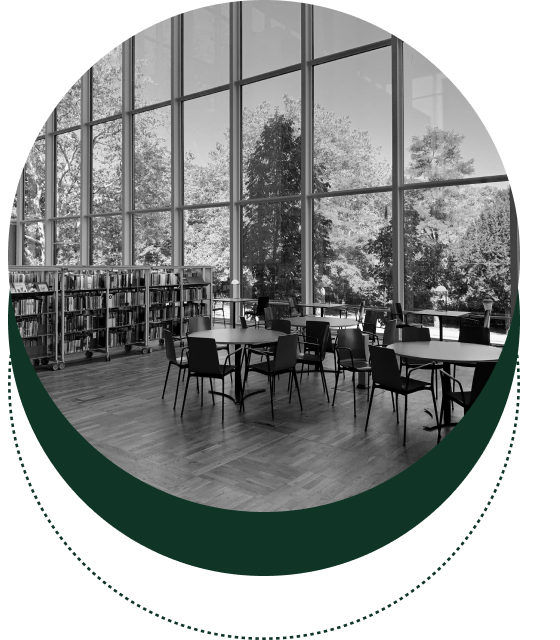 A spacious library with large windows, round tables, and chairs. Bookshelves line the wall. Outside, trees are visible through the windows.