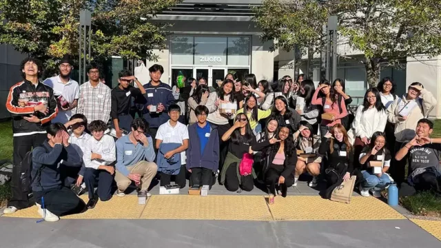 A large group of people, mostly young adults, stand and sit together smiling for a group photo outdoors in front of a building with a sign that reads "Zulora." This vibrant gathering exemplifies the camaraderie fostered by Zuora Careers.