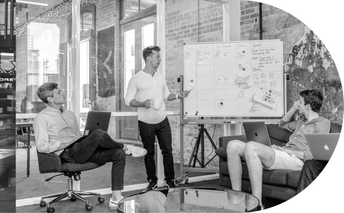 Three men are in a modern office; one is standing and pointing at a whiteboard with charts and notes, while the other two sit, one with a laptop and the other with a notepad.