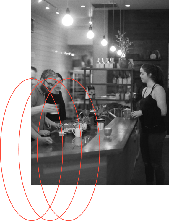 A person behind a counter is preparing a beverage, while another person is standing on the other side of the counter, in a well-lit cafe setting.