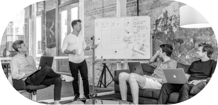 A man standing and pointing at a whiteboard speaks to three seated men with laptops in a casual office setting.