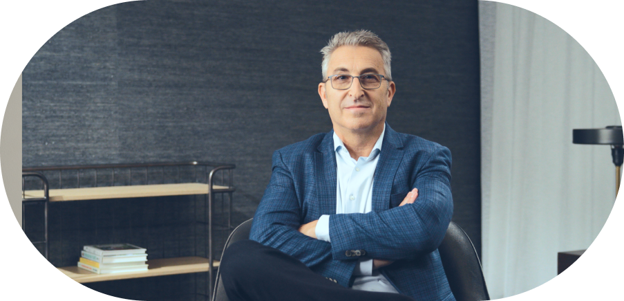 A person with gray hair and glasses, dressed in a blue blazer, sits with arms crossed in an office setting. A bookshelf with stacked books and a lamp are visible in the background.