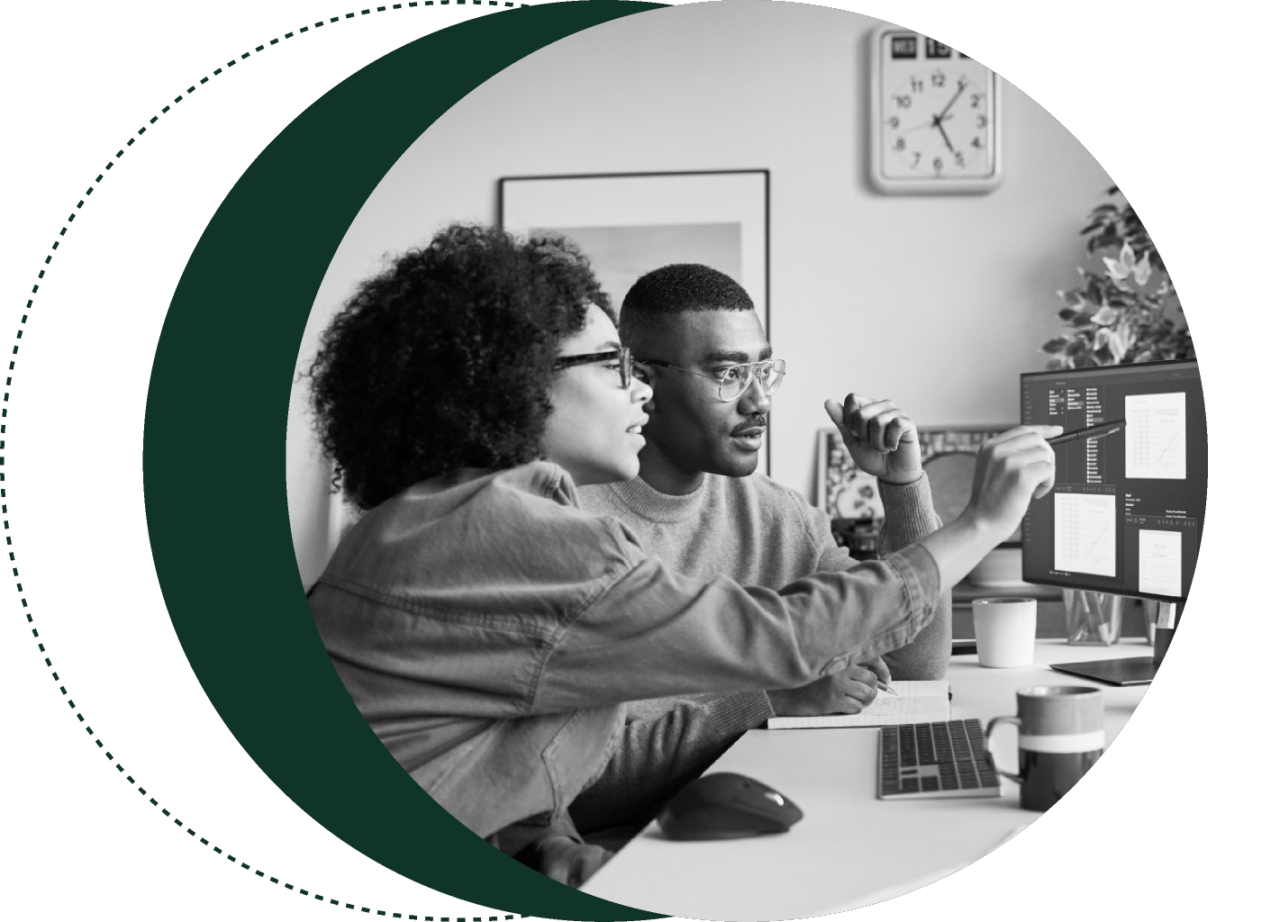 Two people in an office setting analyzing data on computer screens. One person is pointing at the screen while both wear glasses and focus on the content. A clock and office supplies are in the background.