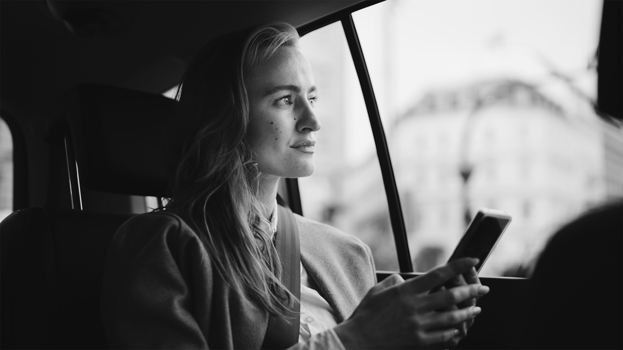 A woman sitting in a car, looking out the window with a thoughtful expression, holding a smartphone in her hand. The photo is in black and white.