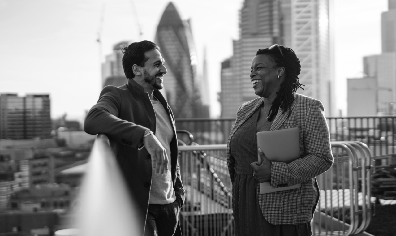 Two people stand on a rooftop, smiling and talking, with a city skyline, including distinctive modern buildings in the background. One person holds a laptop. Black and white photo.