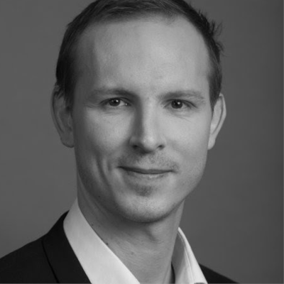 Portrait en noir et blanc d'un homme aux cheveux courts, portant une veste de costume et une chemise blanche, sur un fond uni.