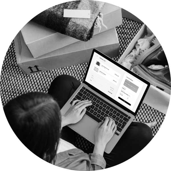 A person sits on the floor with a laptop, shopping online. Boxes and clothes are scattered around.