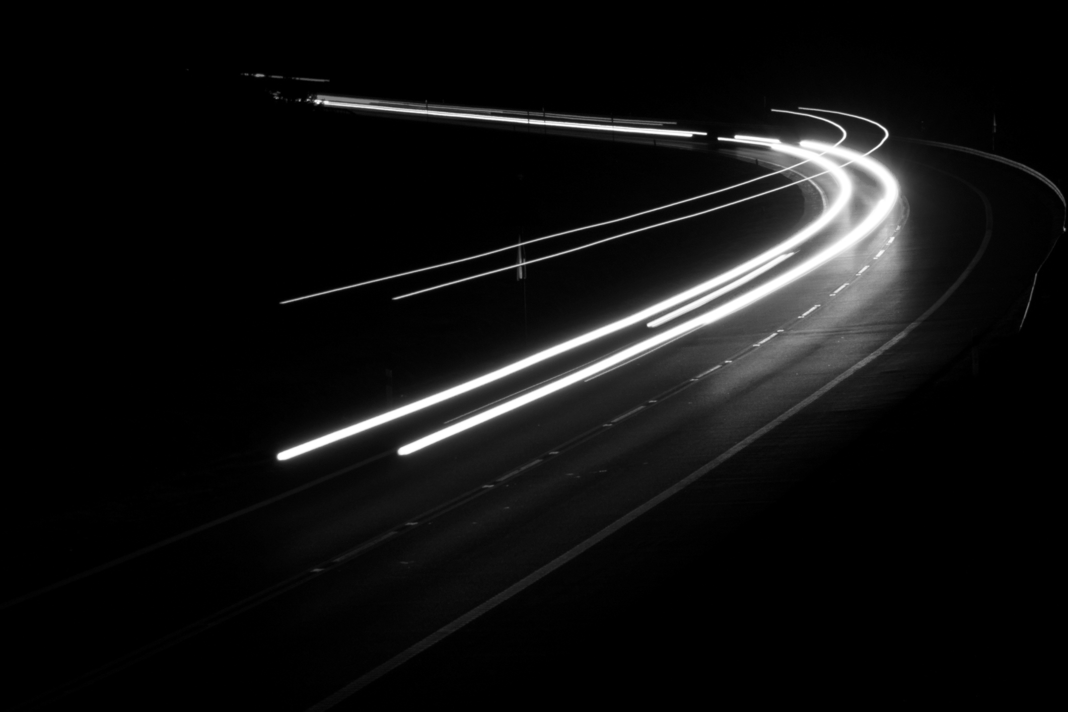 White streaks of light from moving vehicles curve along a dark, empty road at night, captured in a long-exposure photograph.