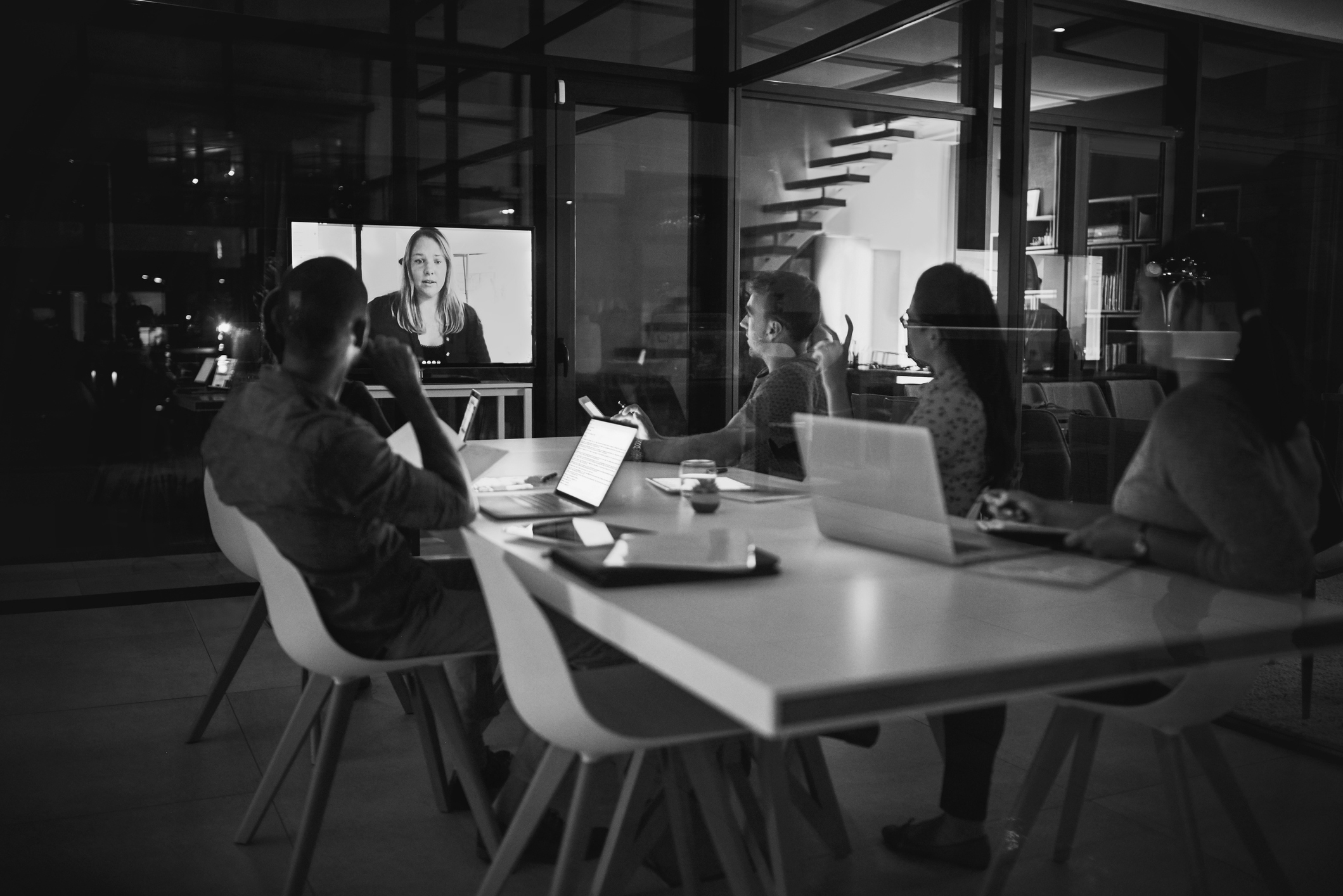 Four people sit at a conference table with laptops, participating in a video call with a woman displayed on a large screen in a modern office setting.