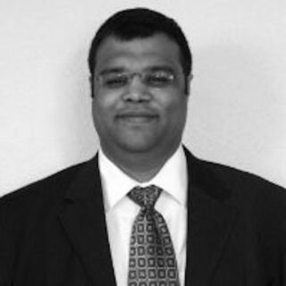 Sid Sanghvi im Anzug und mit Krawatte blickt lächelnd in die Kamera, vor einem schlichten Hintergrund. Das Bild ist in Schwarz-Weiß gehalten.