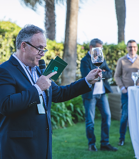 スーツを着た男性がマイクとワイングラスを持って屋外で話しており、その背後では2人が背の高いテーブルの近くに立っている。.