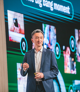 スーツを着た男性がステージ上に立って話している。背後の大きなスクリーンには映像と「ビッグバンの瞬間」というフレーズが映し出されている。.