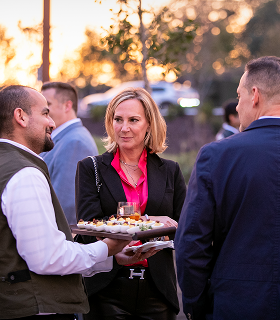 イベントで、サーバーが前菜のトレイを配っている間、ピンクのブラウスを着た女性が屋外に立って2人の男性と話している。.