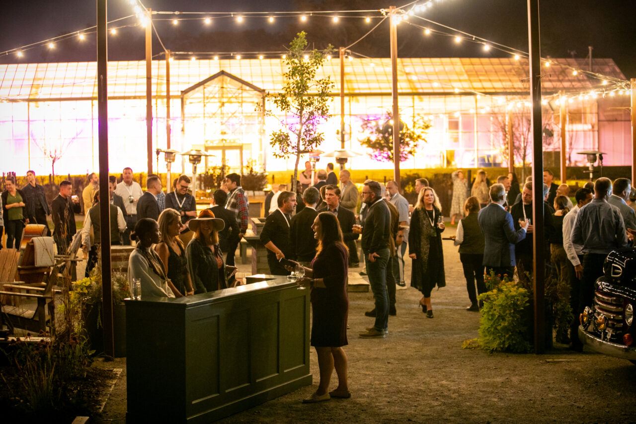 夜になると、人々はバーの近くの電飾の下に屋外に集まり、背景には大きな照明付き温室があります。.