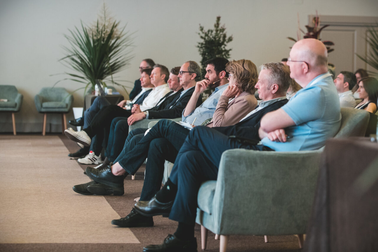 A group of people seated in armchairs attentively watches a presentation or event in a modern, well-lit room with plants in the background.