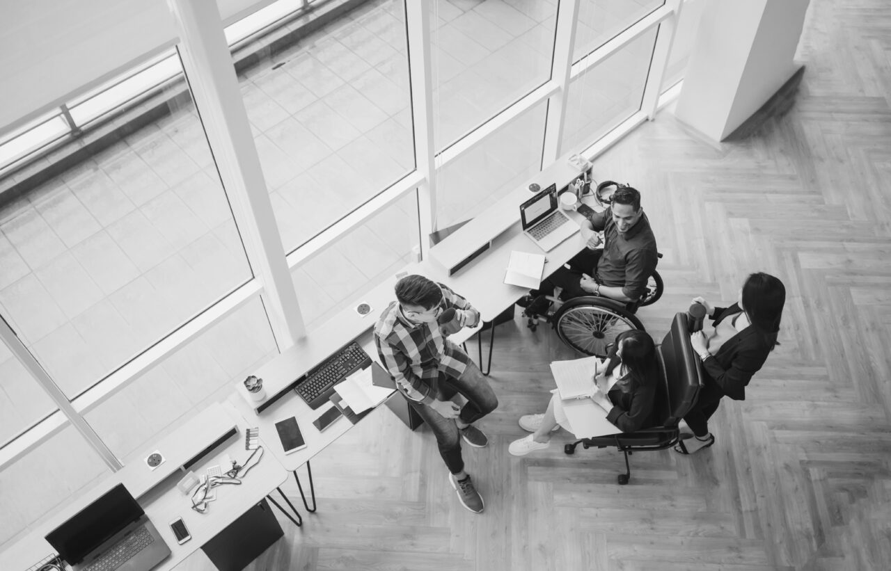 Four people, including one using a wheelchair, have a discussion in a modern office space with large windows and laptops on a long desk.