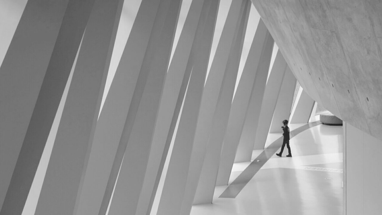 A person walks alone in a modern, spacious interior with large, angled beams and smooth surfaces, captured in black and white.