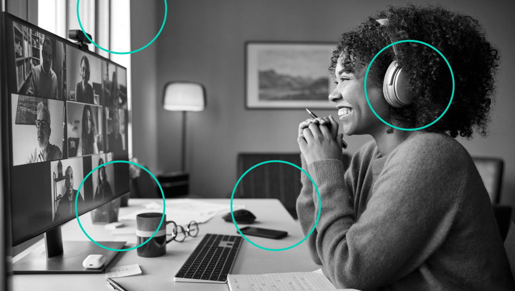 A woman wearing headphones smiles while participating in a video conference with several people on her computer screen in a home office setting.