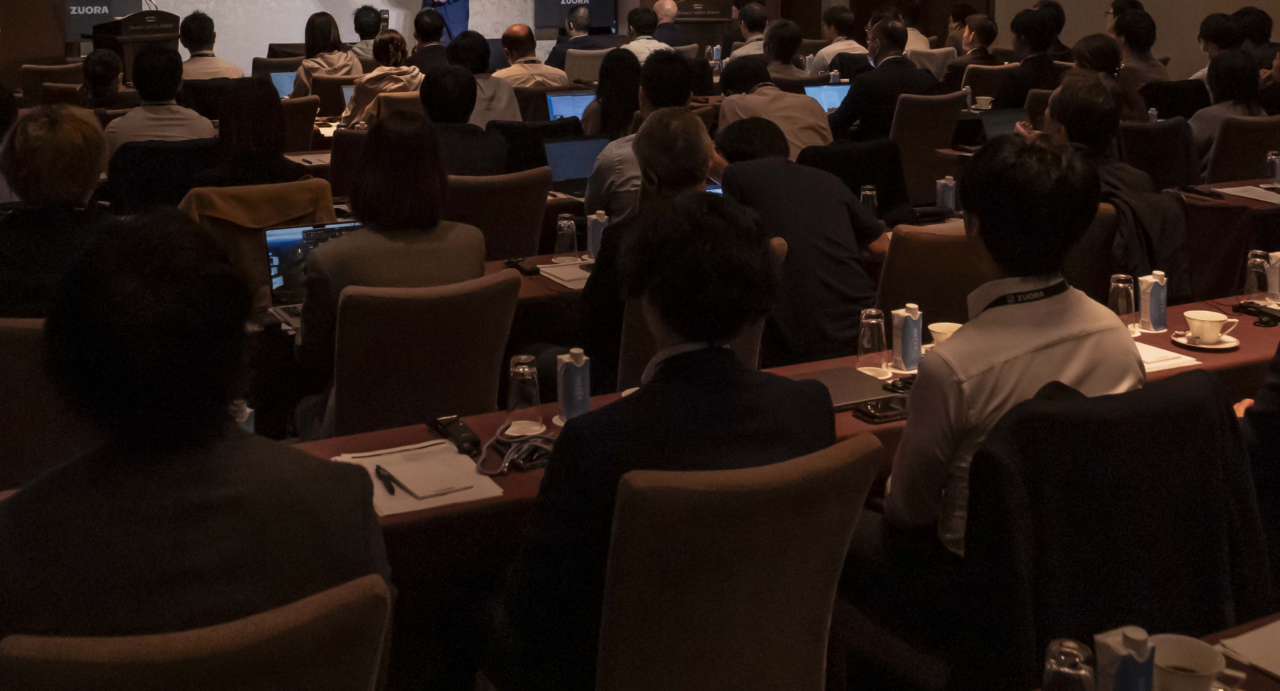 スピーカーが薄暗い会議室で聴衆に向かってスライドを提示し、出席者はテーブルの上にノートパソコン、ノートブック、飲み物を置いている。.