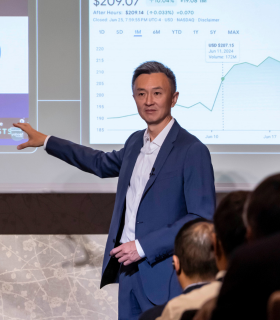 青いスーツに身を包んだ男が、大スクリーンに映し出された金融データを観客に提示する。.