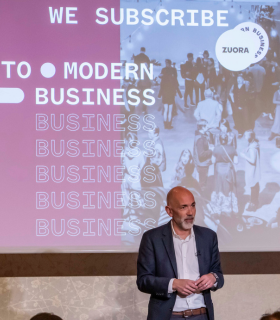 ビジネスイベントで「We Subscribe to Modern Business」の文字と「Zuora」の社名が表示されたスクリーンの前に立ち、スピーチする男性。.