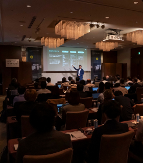 薄暗い照明とシャンデリアの会議室で、青いスーツを着たスピーカーがスライドを聴衆に見せる。.