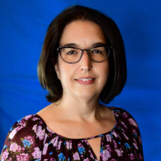 Une femme aux cheveux bruns mi-longs et aux lunettes, portant un chemisier floral violet, pose devant un fond bleu uni.