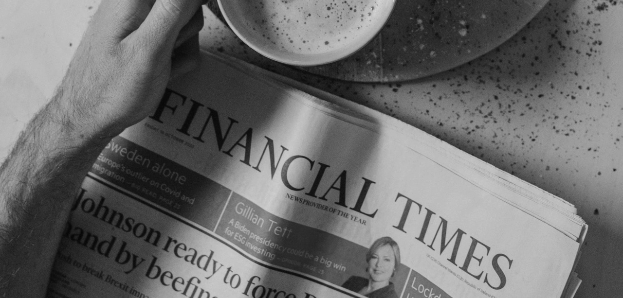 A hand holds a copy of the Financial Times newspaper next to a cup of coffee on a table.