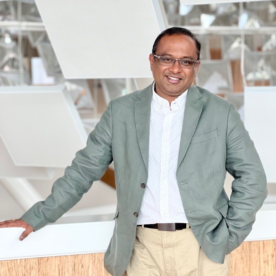 A man wearing glasses, a light green blazer, and beige pants stands indoors, leaning against a white railing with geometric decor in the background.