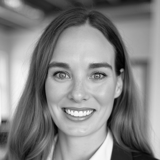 A woman with long hair smiles at the camera in a professional indoor setting, wearing a blazer. The image is in black and white.