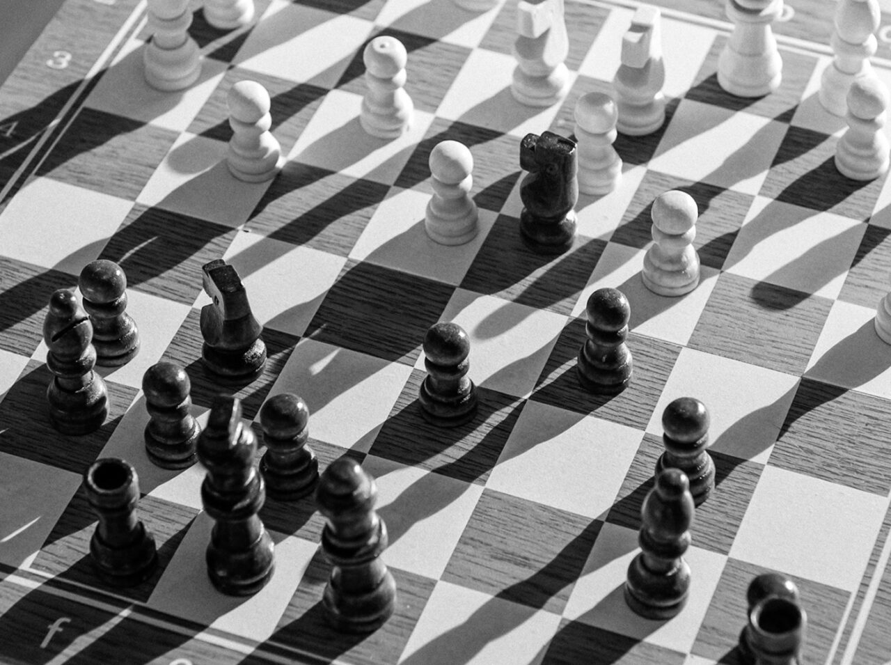 Black and white photo of a chessboard mid-game, evoking the strategy of finance, with both black and white chess pieces casting dramatic shadows across the board.
