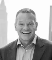 A man in a suit jacket and collared shirt smiles at the camera with a blurred cityscape in the background.