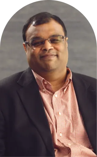 A man wearing glasses, a black blazer, and a red checkered shirt sits in front of a blurred gray background.