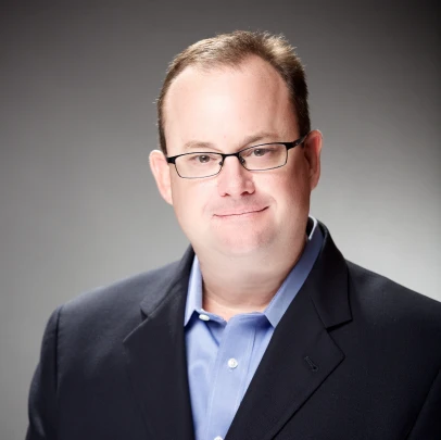 A man wearing glasses, a blue shirt, and a dark blazer poses against a plain, softly lit background.