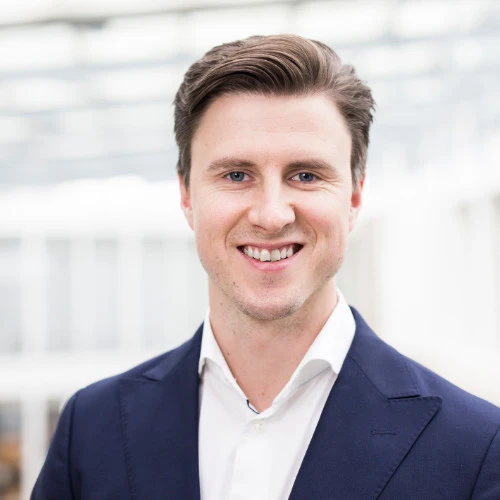 A man with short brown hair wearing a navy suit jacket and white shirt smiles at the camera against a bright, blurred indoor background.