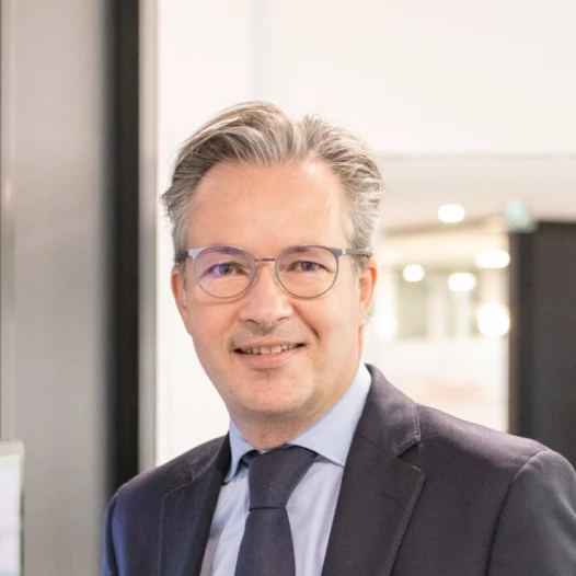 Un homme aux cheveux gris et aux lunettes, vêtu d'un costume sombre, d'une chemise bleu clair et d'une cravate marine, se tient à l'intérieur d'un bureau moderne et sourit à l'appareil photo.