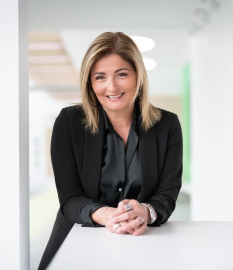 Une femme aux cheveux blonds, vêtue d'un blazer noir et d'une chemise sombre, sourit en s'appuyant sur une surface blanche dans un intérieur moderne et lumineux.