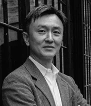 A man in a suit jacket and collared shirt stands in front of a metal fence, looking at the camera with arms crossed.