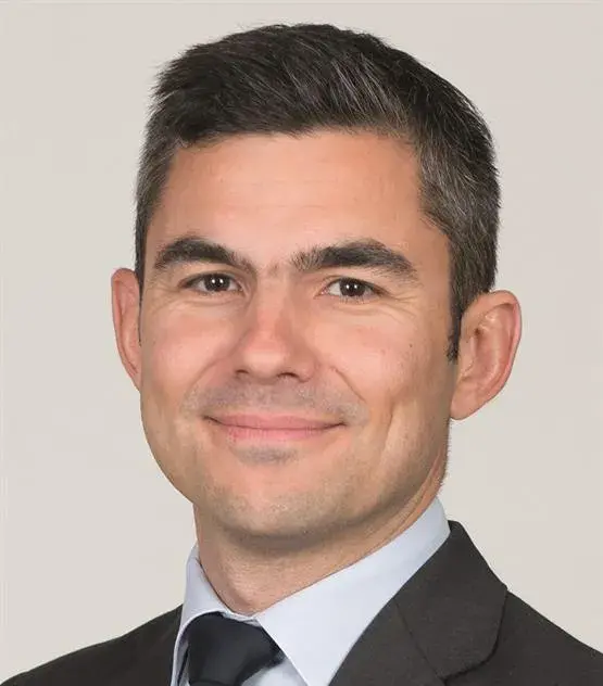 Un homme aux cheveux bruns courts et à la barbe légère, portant une veste de costume sombre, une chemise claire et une cravate sombre, pose sur un fond uni.