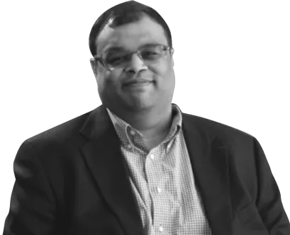 A man wearing glasses, a blazer, and a checkered shirt is looking at the camera and smiling in a posed portrait.