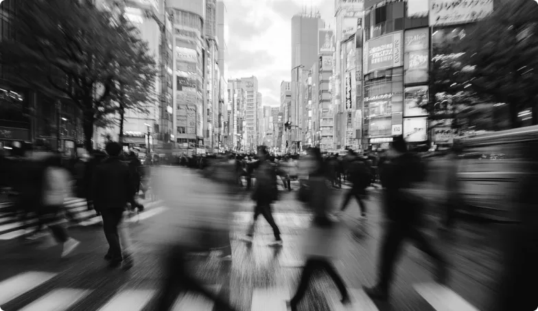 Black and white photo of people crossing a busy city street with tall buildings and bright signs in the background; motion blur captures movement.