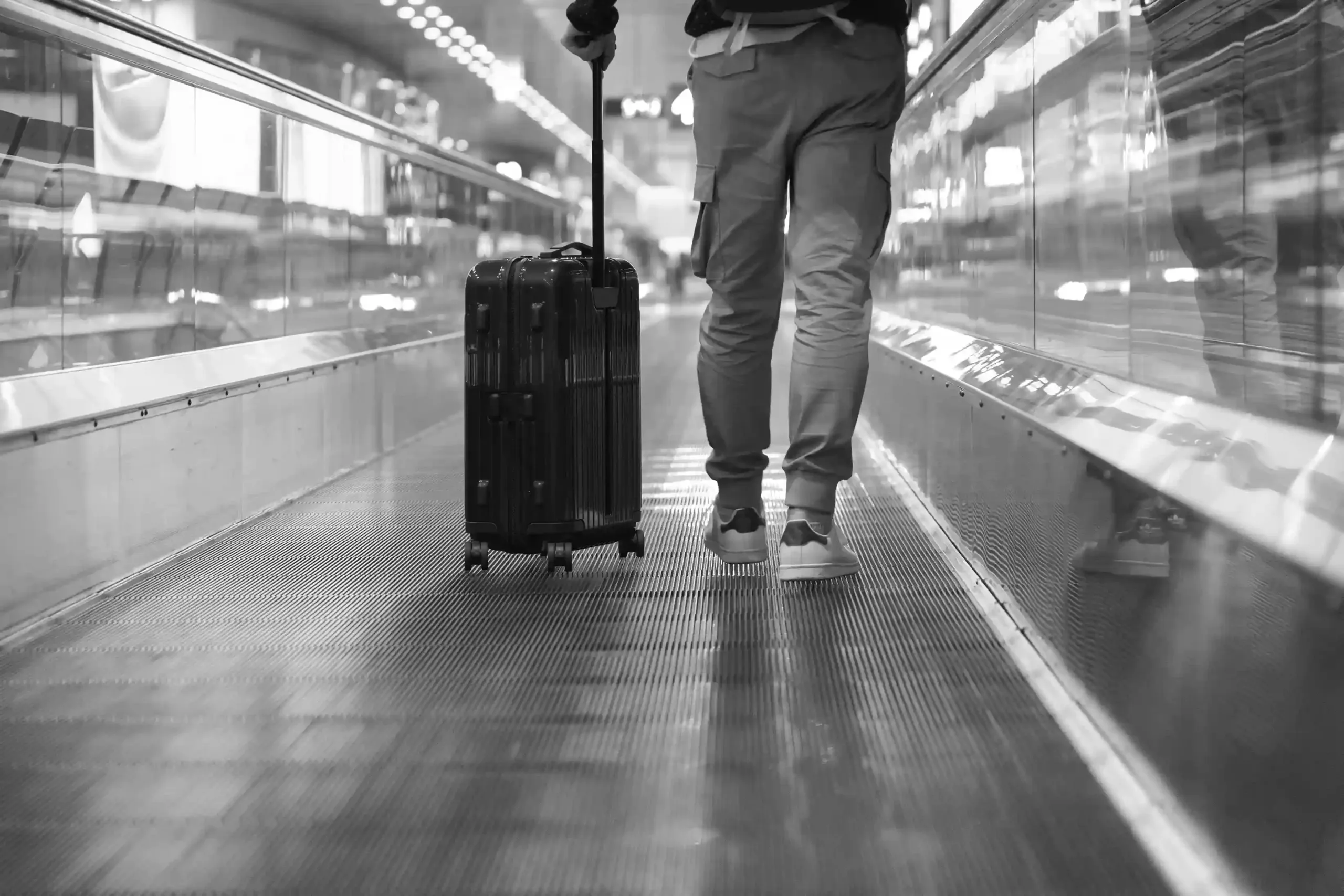 Person walking on a moving walkway in an airport, pulling a wheeled suitcase behind them. The image is in black and white.